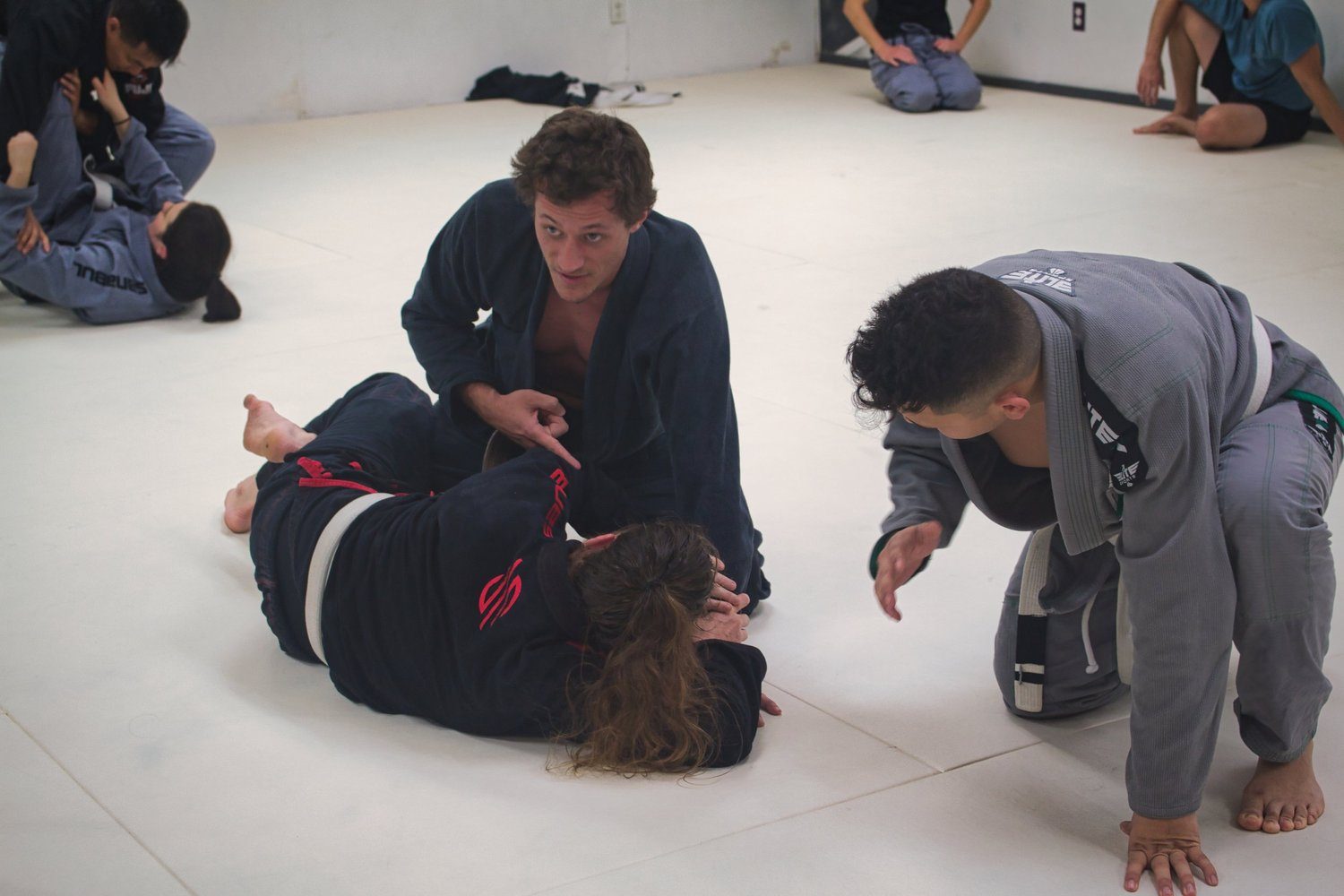 Instructor kneeling between two pairs of students on the mats to explain a grappling detail.