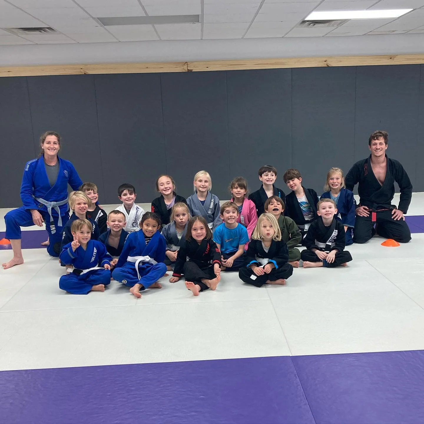 Homeschool and youth jiu-jitsu students smiling on the purple and white mats with Coach John and Coach Sarah.