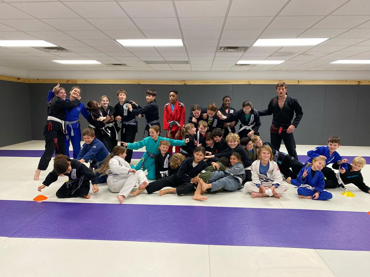 Large group of kids and teens laughing on the purple and white mats after class with their coaches.