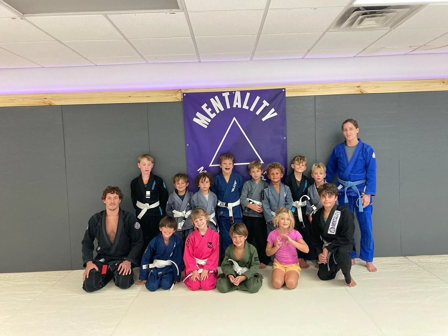Coach John and Coach Sarah on the mats with the youth jiu-jitsu class in front of the Mentality banner.