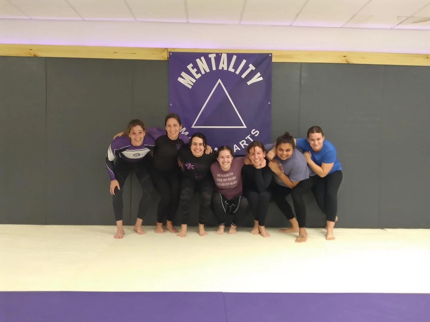 Seven women smiling together on the mats in front of the Mentality Martial Arts banner.